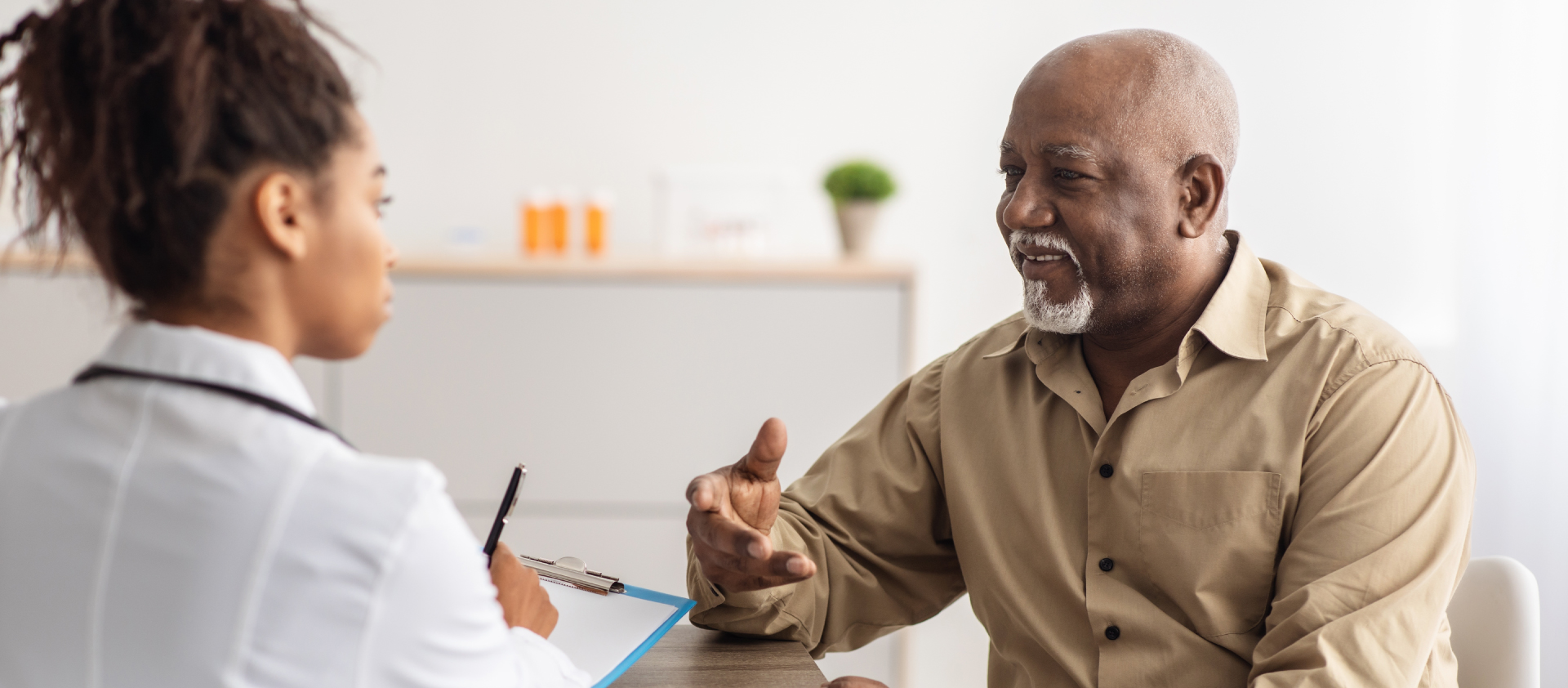 a female doctor holding a clipboard talks with a smiling older male patient