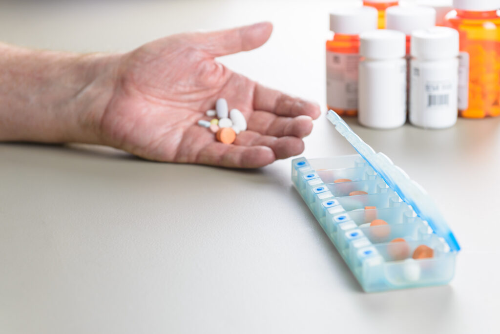 A person holding several different pills in their hand, and a pill organizer and multiple medication bottles in the background.